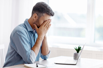 Naklejka premium Remote Work Stress. Tired freelancer man sitting at desk with laptop at home office, overworked man covering face with hands, feeling fatigue and lack of inspiration, having problems with project