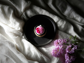 Elegant dessert on a black plate surrounded by white fabric and lilac flowers