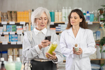 Mature woman in pharmacy scans QR code on shampoo product, checks certificate on manufacturers website and consulting with woman pharmacist about using cosmetic care product