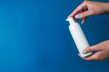 Close-up of two hands holding a white dispenser bottle on a blue background with space for text. The concept of foam cleansers, skin cleansers.