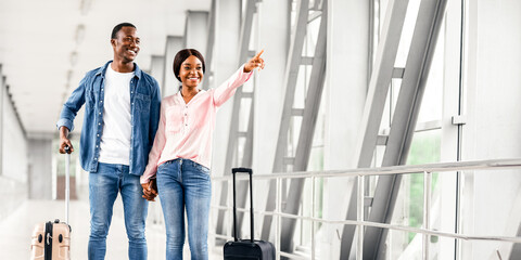 Best Airlines Concept. Happy Black Couple In Airport Pointing Away At Copy Space, Young African American Spouses Walking With Suitcase Luggage In Terminal, Ready For Honeymoon Vacation Trip