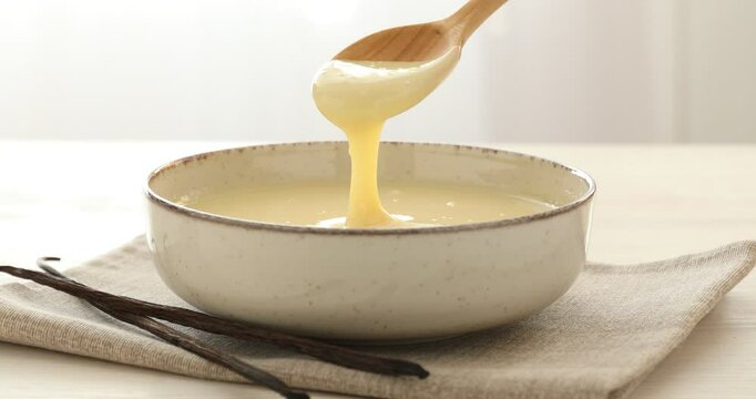 Woman stirring condensed vanilla milk in bowl at light table, closeup