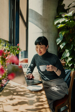 Asian freelancer woman working remotely from coffee shop using laptop and drinking coffee