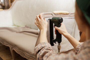 Hands of upholsterer stapling fabric on antique couch with pneumatic stapler in workshop.