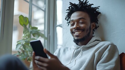 Young African American man with dreadlocks smiling while using smartphone at home, wearing light gray hoodie. Natural window lighting and indoor plant create cozy atmosphere.
