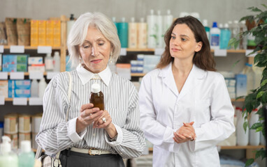Obraz premium Mature woman choose expectorant syrup, buys product in drug store. Checking for absence of GMOs, age restrictions and recommendations for use. Woman pharmacist offers product, promotes product of