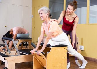 Personal female trainer controlling movements of senior woman doing pilates on combo chair in fitness studio. Healthy active lifestyle