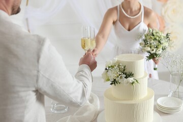 Married couple clinking glasses of champagne at table with wedding cake indoors, closeup