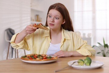 Sad teenage girl with pizza and broccoli at wooden table indoors. Eating disorder