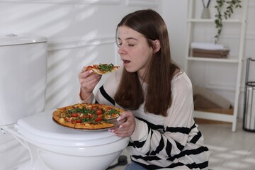 Teen girl eating pizza near toilet bowl at home. Bulimia