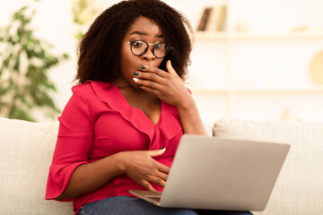 Fototapeta premium No Way, I Can't Believe. Portrait ot surprised african american lady in glasses covering mouth in amazement, reading unexpected news on laptop computer, sitting on the couch at home, copy space
