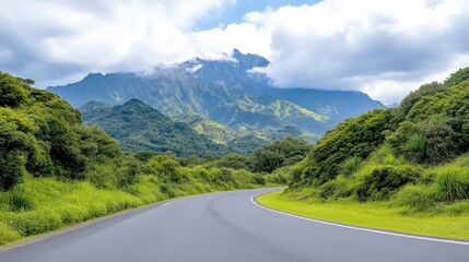 Naklejka premium A winding asphalt road curves gently through lush green hills. Mountains rise in the background, partly shrouded in clouds. The image is high-resolution, showcasing vibrant colors and sharp detail un