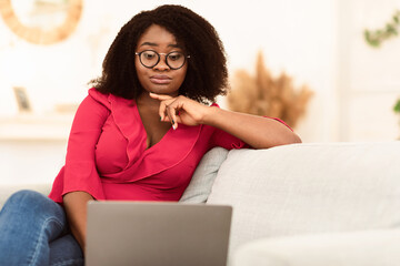 Portrait of confused skeptic black curly woman in eyeglasses looking at her laptop sitting on couch...