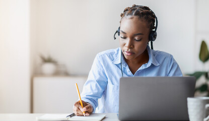 Online Training. Young black female employee in headset watching webinar on laptop and taking...