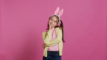 Smiling little girl dreaming about gifts and food for easter sunday celebration, feeling excited about spring holiday. Young kid with bunny ears posing against pink background. Camera B.