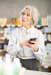 Elderly woman with a mobile phone in her hands chooses medicines in a pharmacy and looks at an...