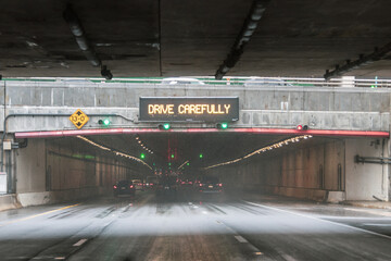 Traffic warning sign advises caution while vehicles navigate through urban underpass during rainy...
