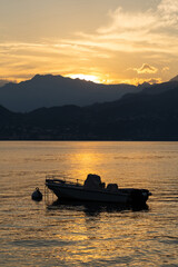Tranquil boat floats on lake during sunset at Varenna, lake Como, Italy, silhouetted against golden sky and mountains in background. Water reflects warm hues of fading sunlight