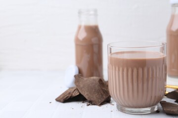 Tasty chocolate milk in glass and pieces of chocolate on white tiled table, closeup. Space for text