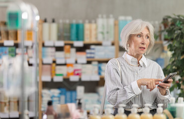 Elderly female shopper searches for necessary medications on shelves of a pharmacy, looking through the list on her smartphone