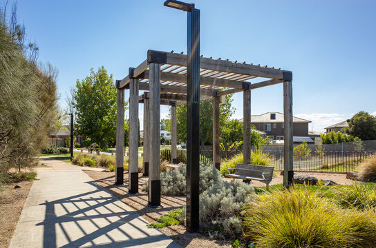 A modern wooden pergola with parallel beams, situated in a public park or communal garden and surrounded by drought-tolerant plants, including ornamental grasses and shrubs, in a suburb of Australia. 