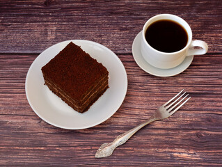 A piece of layered sponge cake with chocolate, a fork and a cup of hot black coffee on a wooden table.