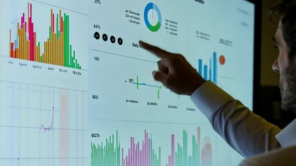 Man analyzes financial data on a large screen, pointing at charts and graphs display