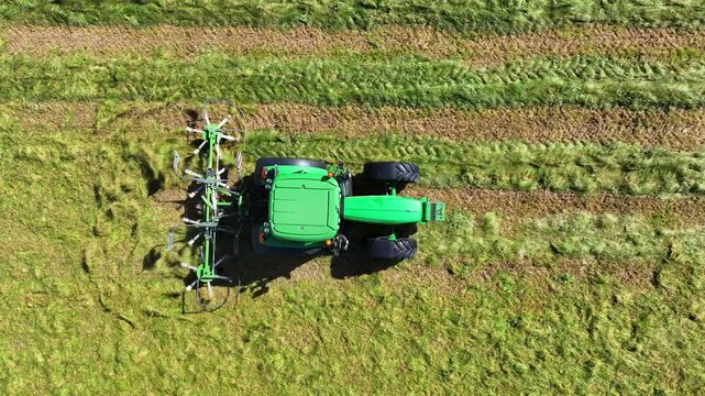 Rear-mounted tedder behind green tractor spread cut hay in farm field, top drone