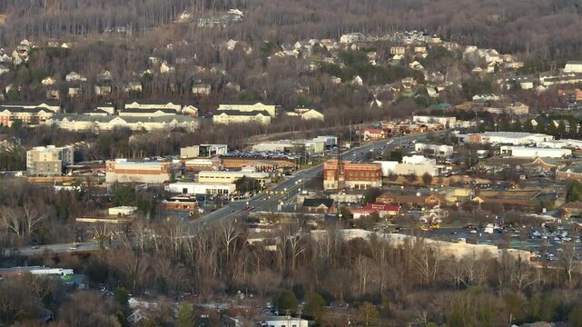Aerial zoom shot of suburb intersection with houses and shops in Charlottesville. Neighborhood located on hill lighting by sunset. Wide shot. Leafless forest trees in spring. VA, USA.