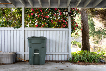 Naklejka premium Empty carport with yard waste bin, camellia bush in background full of pink flowers is full bloom, sunny spring day 