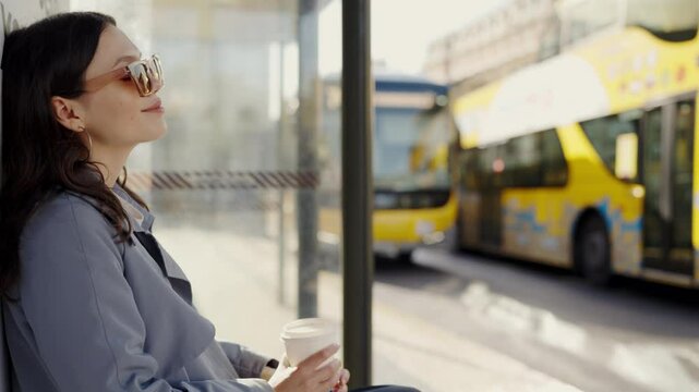 A young woman in sunglasses sips coffee and smiles, sitting at a bus station on a sunny day. Blurred yellow bus in the background. Young Woman Enjoying Coffee While Waiting at Bus Station