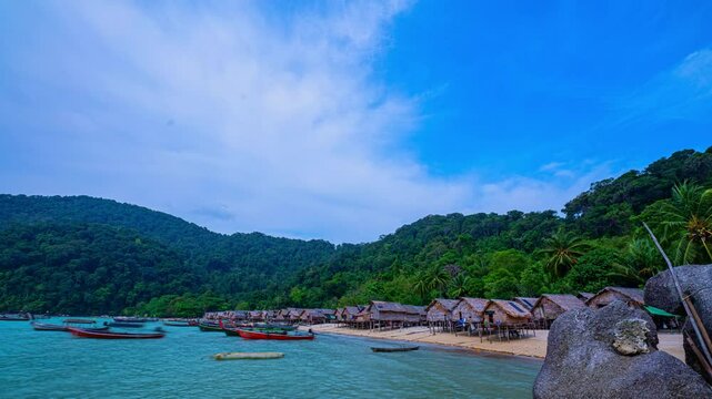 Time lapse a tropical beach village with rows of small huts along the coastline, surrounded by lush green jungle. Traditional fishing boats are anchored in the turquoise waters, creating a vibrant