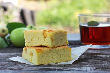 A slice of charlotte pie rests on a wooden table against a green apple and a glass of tea. Home baking on a summer afternoon in the garden