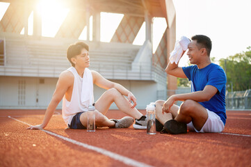 Two asian athlete men resting and wiping sweat, sitting together on racetrack. Jogging or Running.