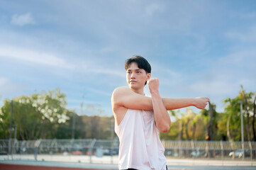 Asian man in white tank top stretching his arm and shoulder muscles on racetrack. Outdoor exercise.