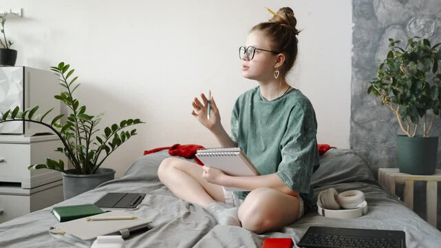 Student immersed in studying, writing in planner while lying on bed. Markers, notebooks, and gadgets scattered around create organized study space. Comfortable home education for effective learning.