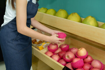 Caucasian woman wearing denim apron arranging apples wrapped in pink netting on wooden shelf in...