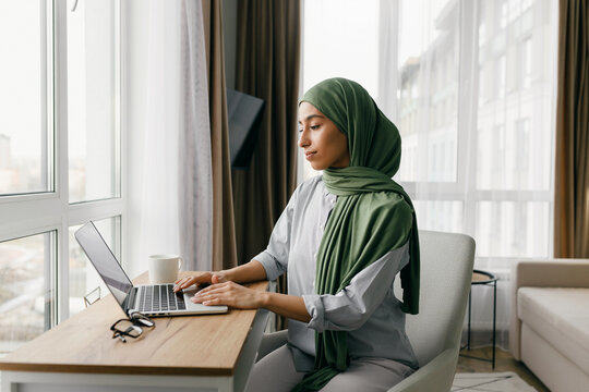 Muslim businesswoman in green hijab using laptop while sitting at wooden desk in cozy home office, working or studying online from home, running small business, selling goods at marketplaces