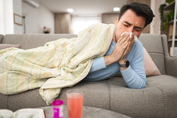 Sick man having a runny nose while lying on a sofa covered with blanket