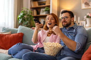 A smiling couple relaxes on the couch, watching scary movies and sharing a bowl of popcorn. Their...