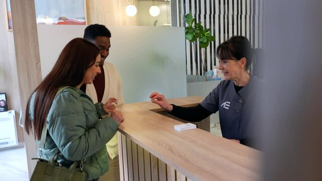 Friendly dental clinic receptionist handing an appointment reminder card to a smiling young couple