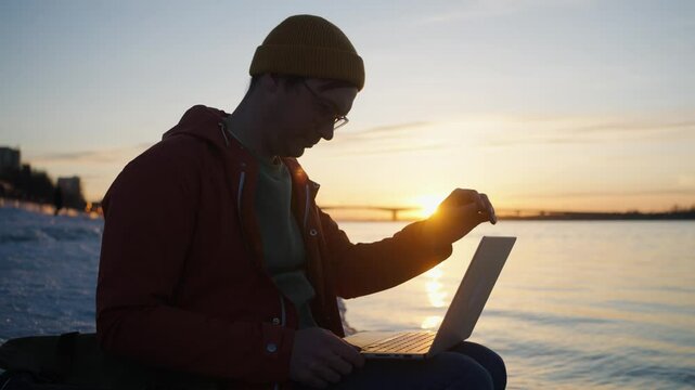 Young freelancer working on laptop by river during golden sunset, embodying digital nomad lifestyle with serene landscape background and peaceful work environment