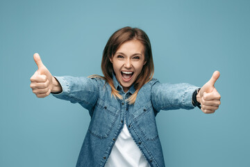 Cheerful young woman in jeans shirt showing thumbs up over blue background. Look in camera