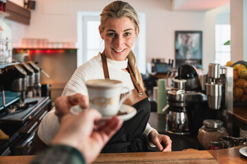 Barista with blonde hair offers a cup of coffee to a guest inside a German café
