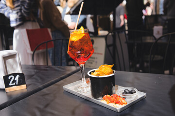 Spritz veneziano cocktail, served with chips, olives, and bruschetta, sits on a table at an outdoor...