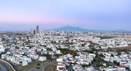 Obraz premium A panoramic view of modern Puebla, Mexico, featuring residential neighborhoods, contemporary high-rise buildings, and a mountainous backdrop under a pastel-colored sky at dawn