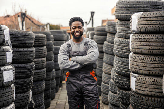Portrait of multicultural auto mechanic worker standing near piles of tires with arms crossed and smiling at camera. - Powered by Adobe