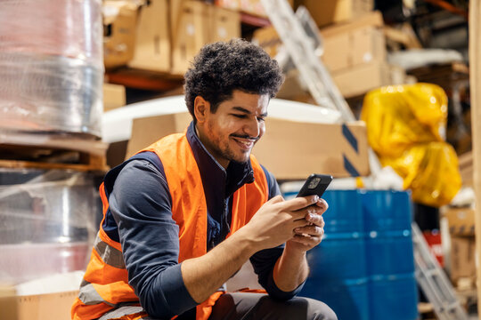 Smiling interracial warehouseman sitting in storage and typing online messages on cellphone.