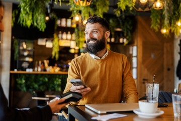 Smiling young bearded businessman sitting in coffee shop and paying bill with his cellphone on pos...