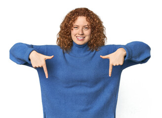 Young Caucasian redhead woman points down with fingers, positive feeling. © Asier